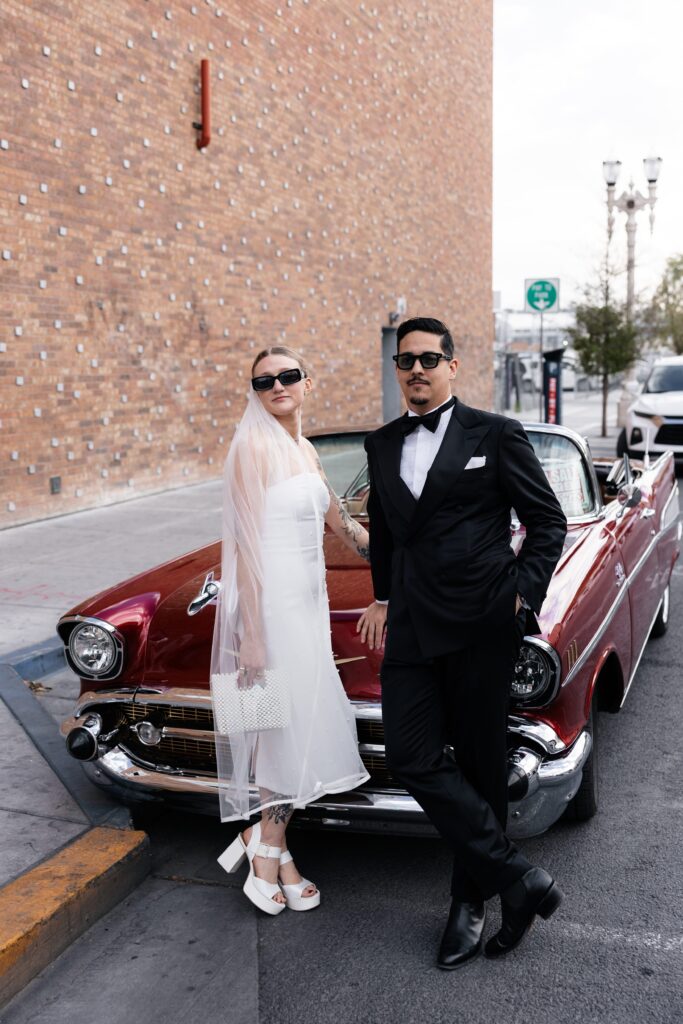 Bride and groom wearing sunglasses as they pose in front of a classic cherry red Chevrolet for their Las Vegas elopement wedding portraits