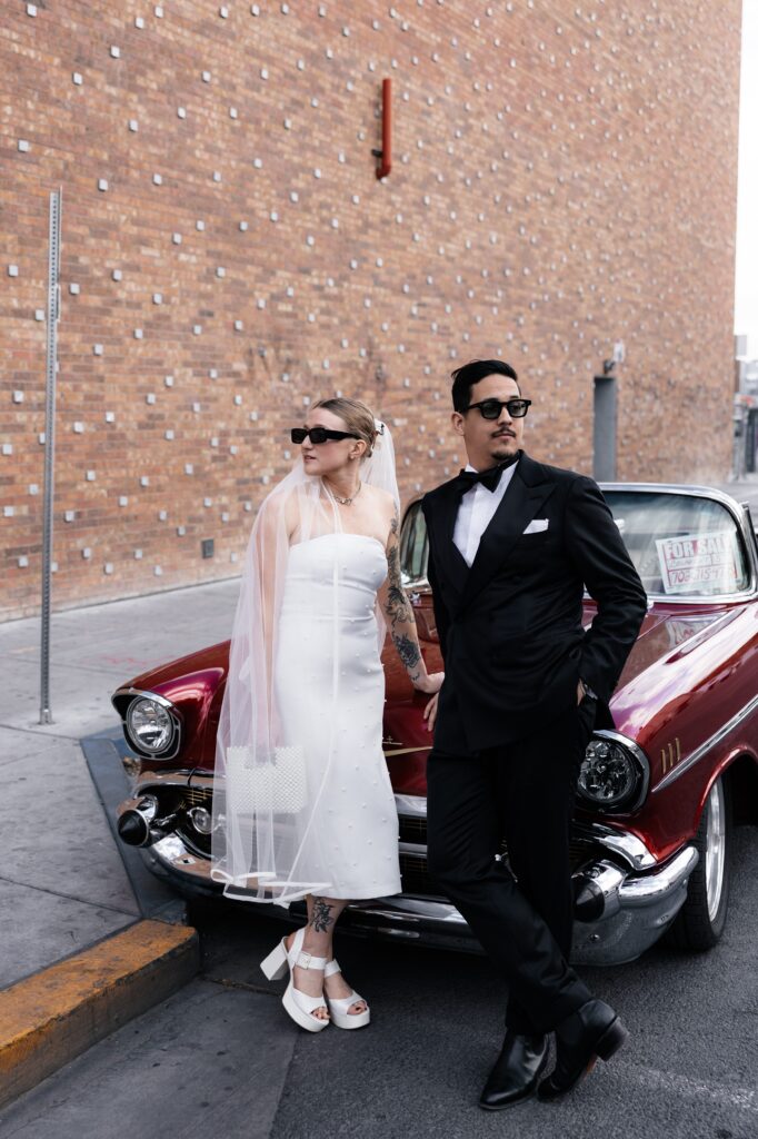 Bride and groom wearing sunglasses as they pose in front of a classic cherry red Chevrolet for their Las Vegas elopement wedding portraits