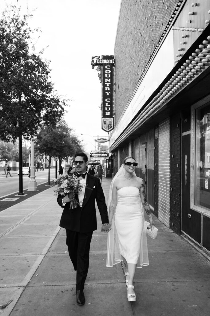 Black and white photo of a bride and groom walking down Fremont Street  wearing sunglasses