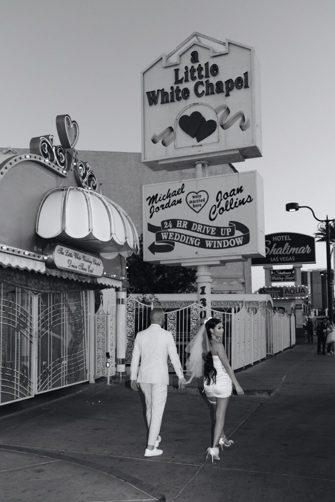 Black and white photo of a bride and groom walking in front of The Little White Wedding Chapel sign
