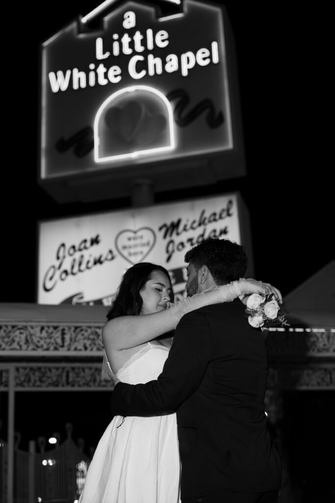 Black and white elopement photo of a bride and groom in front of a Little White Chapel sign