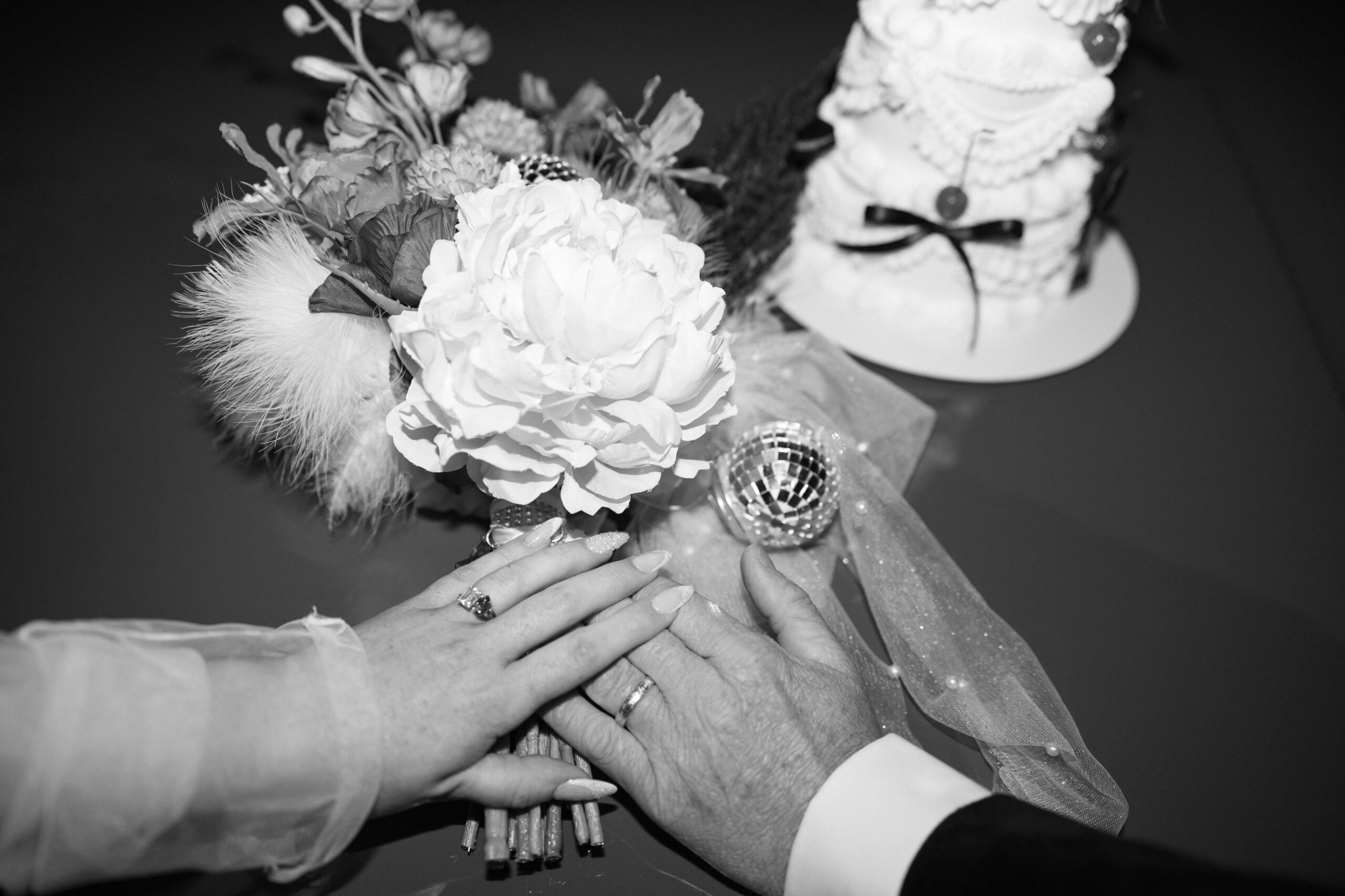 Black and white photo of a bride and grooms hands on top of each others with florals and an elopement cake