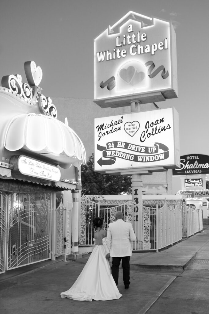 Black and white elopement photo of a bride and groom posing in front of a Little White Chapel sign