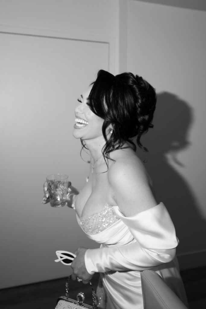 Black and white candid wedding photo of a bride laughing while holding a glass of alcohol, her purse and sunnies. 