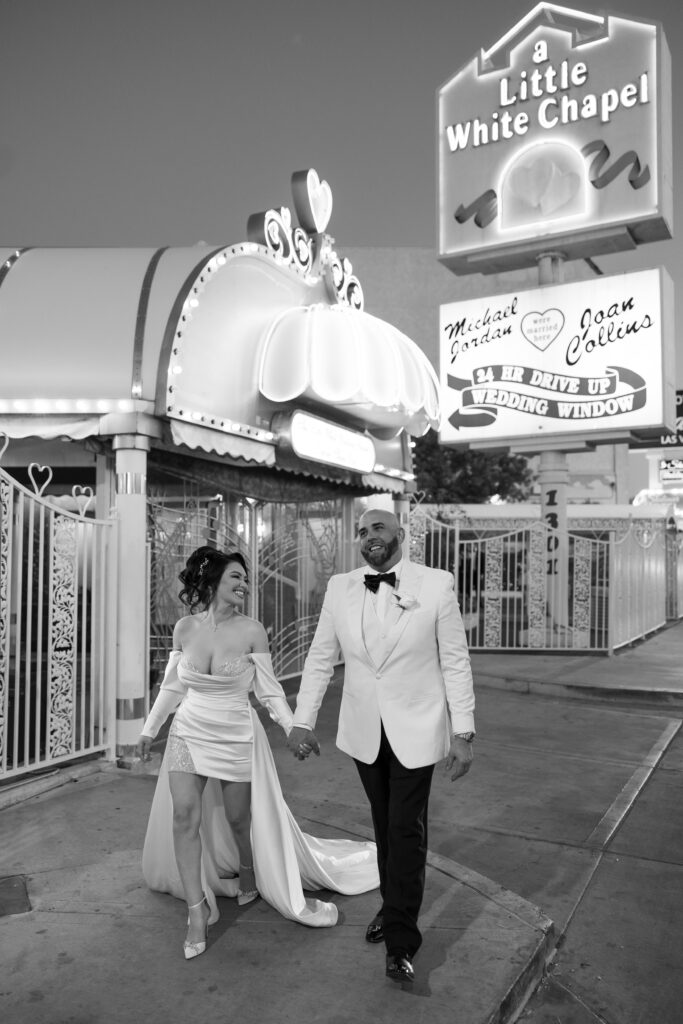 Black and white photo of a bride and groom holding hands as they walk in front of The Little White Wedding Chapel