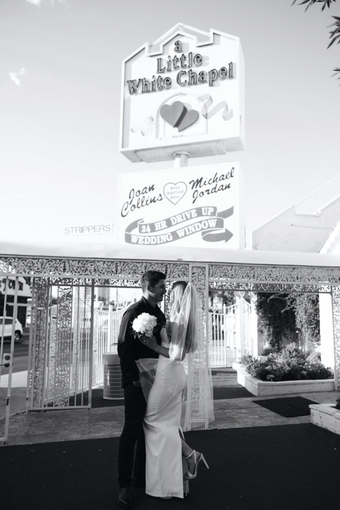 Black and white photo of a bride and groom posing in front of the Little White Chapel sign