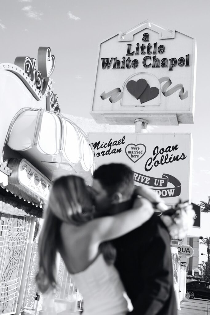 Black and white photo of a bride and groom kissing in front of a Little White Chapel sign