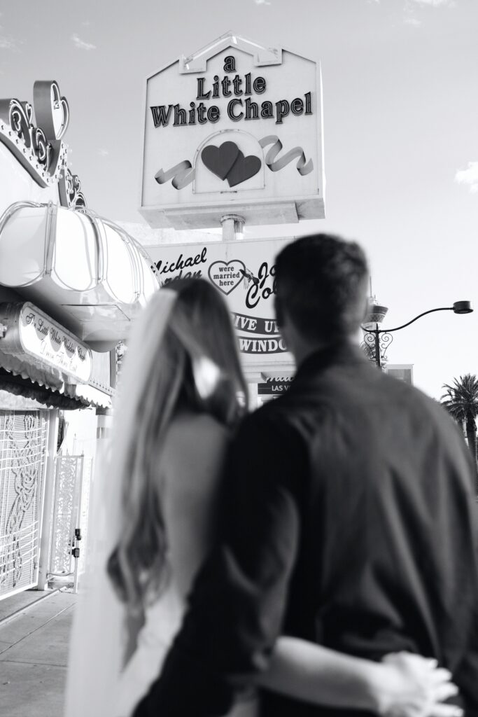 Black and white photo of a bride and groom looking at a Little White Chapel sign