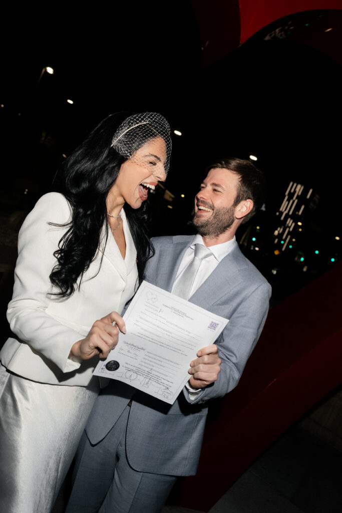 Bride and groom posing with their marriage license outside of the Clark County Marriage License Bureau in Las Vegas at night.