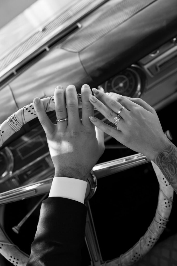 Black and white photo of a bride and groom holding onto the steering wheel during their Pink Cadillac elopement