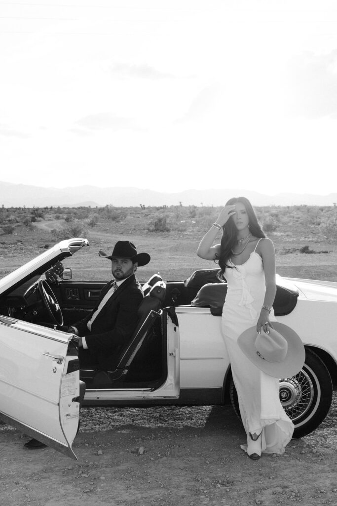 Stylish black and white photo of a couple posing with an open car door during their Las Vegas engagement shoot in the desert.