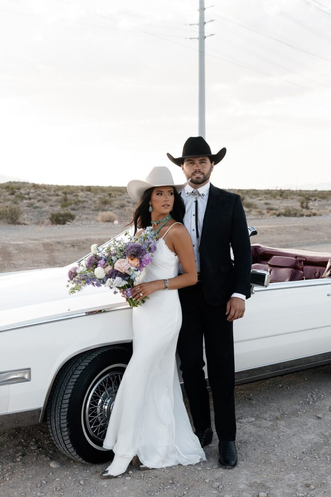 Western engagement photos in Las Vegas featuring a couple in cowboy hats standing beside a white Cadillac with a colorful bouquet in hand.