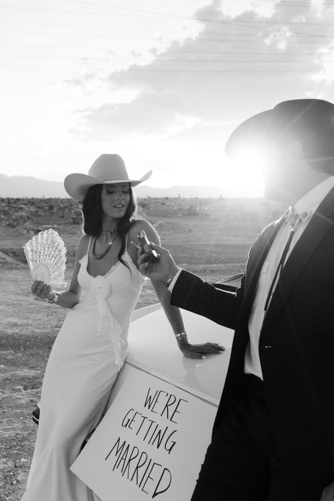 Black and white Las Vegas wedding announcement photo of a couple posing with a car and a "We're Getting Married" sign on the back