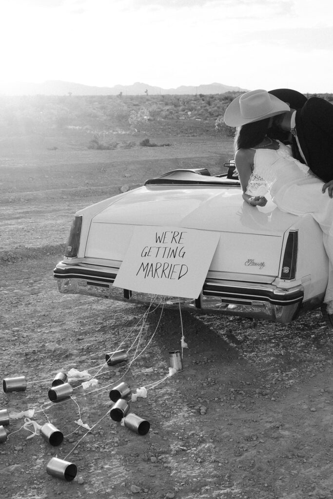 Black and white Las Vegas wedding announcement photo of a couple posing with a car and a "We're Getting Married" sign on the back