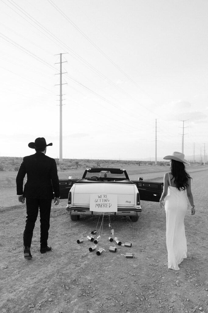 Black and white photo of a couple walking towards a car with a "We're Getting Married" sign on the back