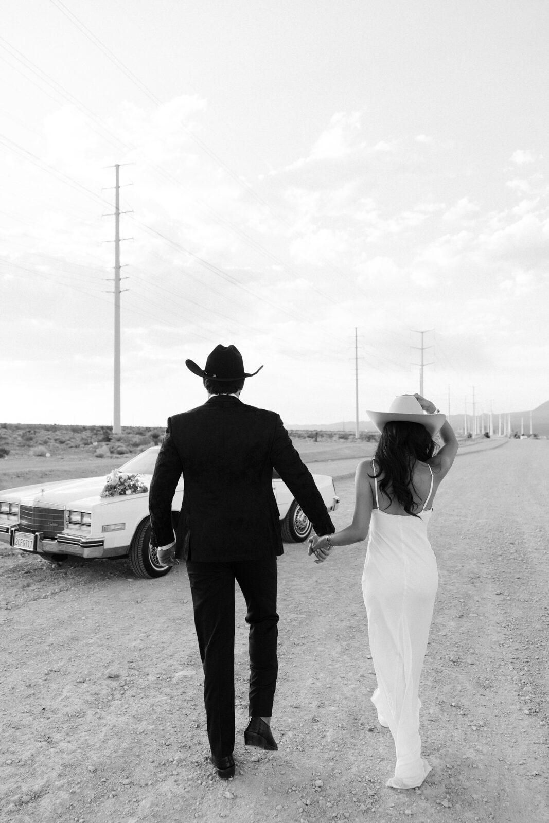 Black and white photo of western engagement photos in Las Vegas with the couple walking toward a vintage Cadillac in the desert.