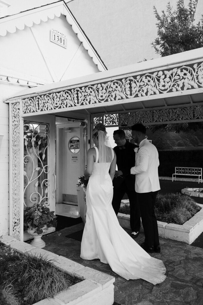 Black and white candid photo of a bride and groom walking into The Little White Wedding Chapel with their officiant. 