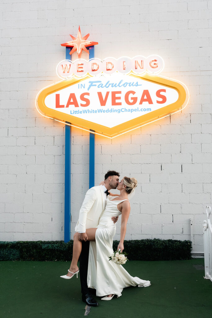 Bride and groom kissing in front a "Wedding in Fabulous Las Vegas" sign outside of The Little White Wedding Chapel.