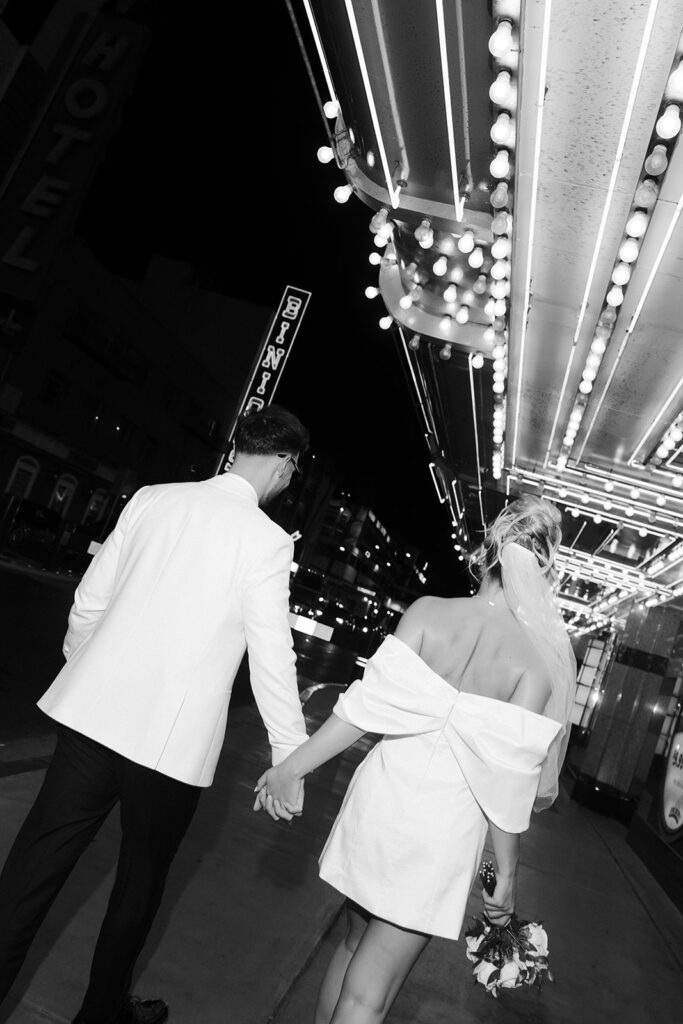 Black and white photo of a bride and groom walking down Fremont Street hand in hand.