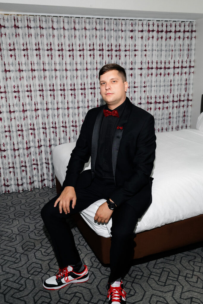 Groom sits on a hotel bed wearing a black suit and red sneakers during getting ready at a Cosmopolitan wedding in Las Vegas.