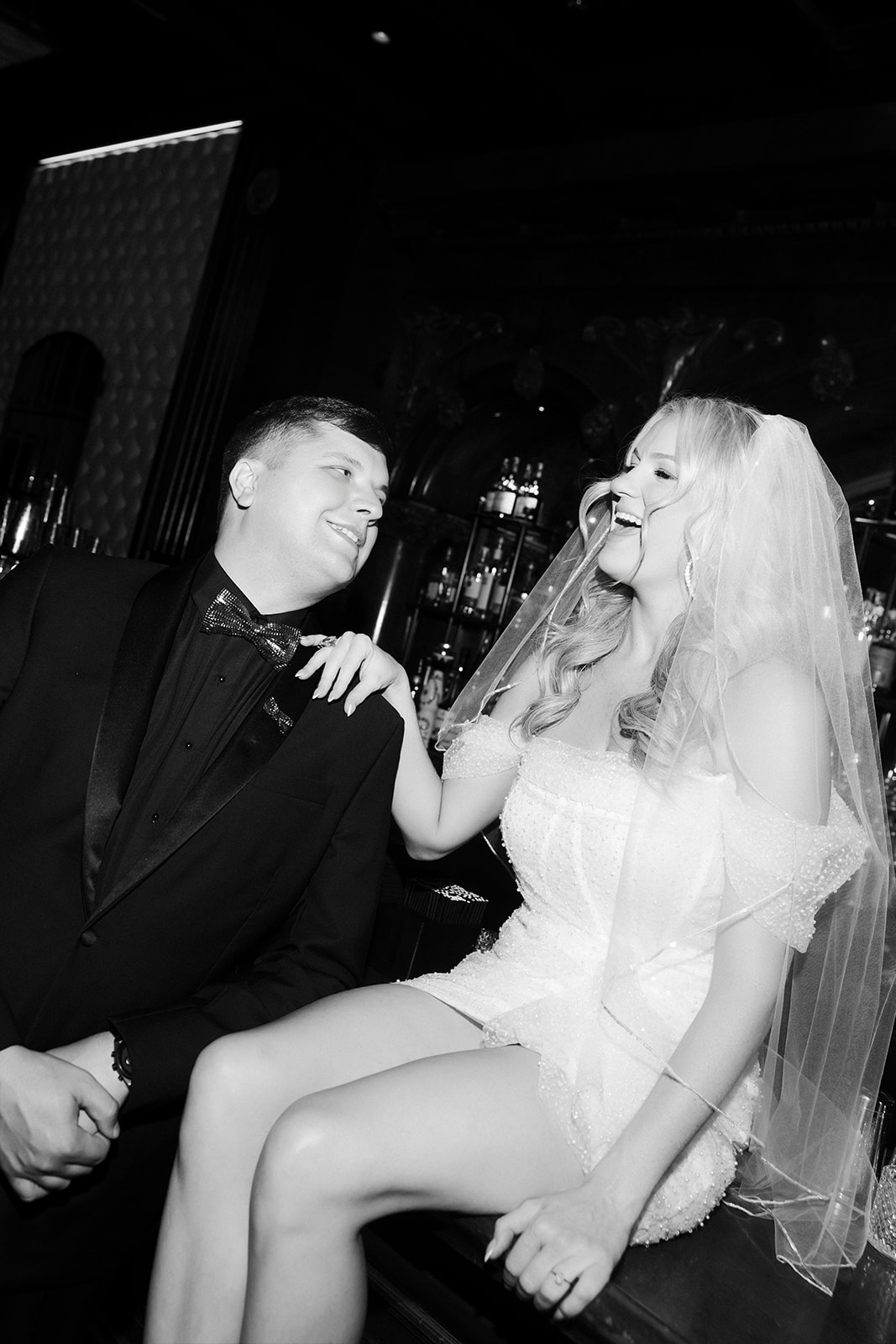 Bride laughing while sitting on a bar counter next to the groom during black-and-white Las Vegas wedding portraits.