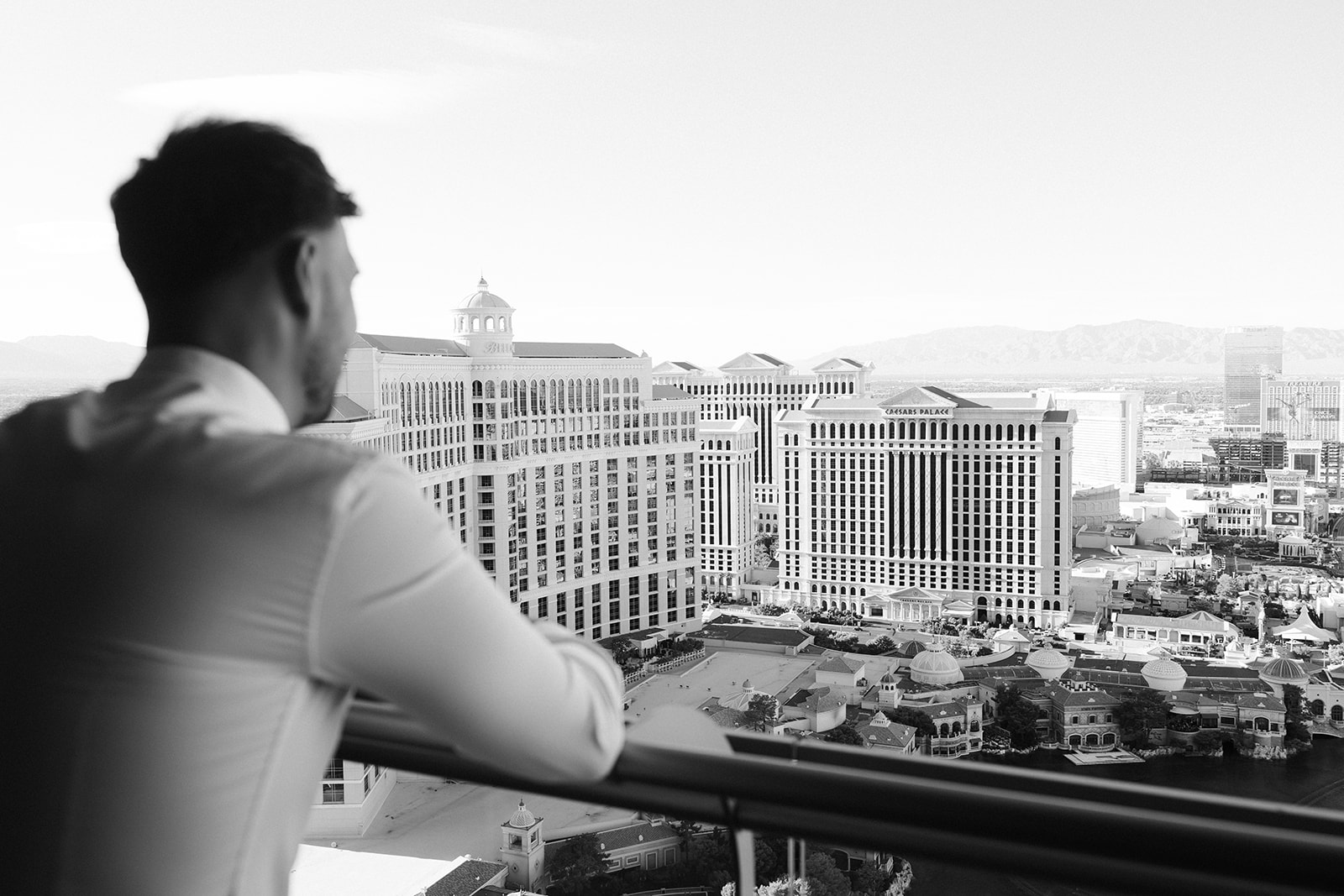 Black and white wide shot of a groom looking out at the city of Las Vegas from the balcony at The Cosmopolitan.