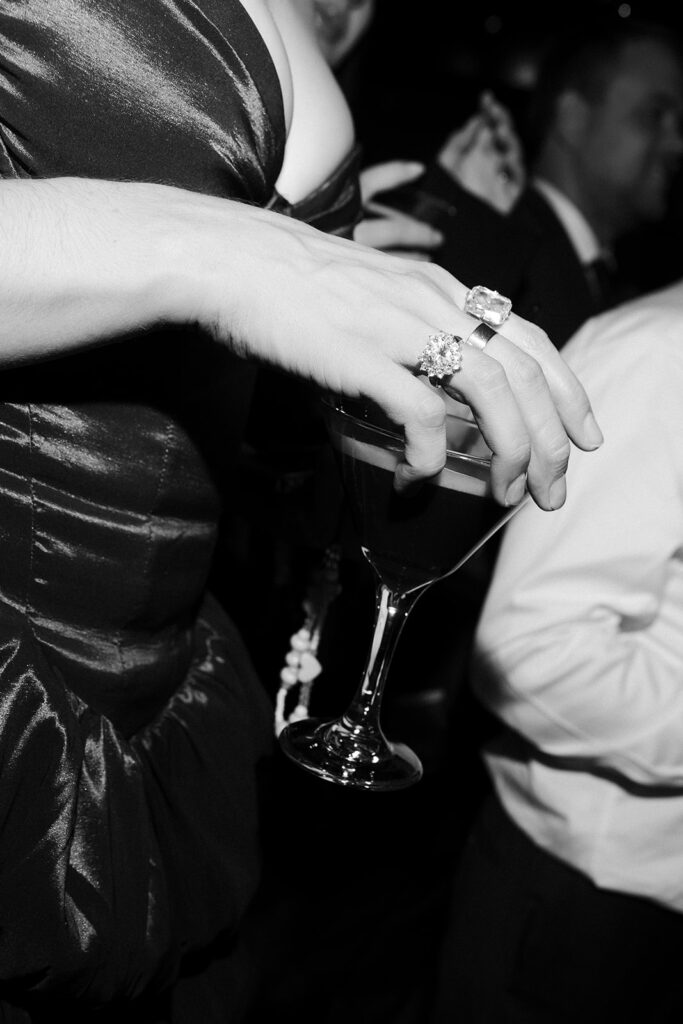 Black and white close-up of a guests hand holding a cocktail glass, highlighting her rings during the reception.