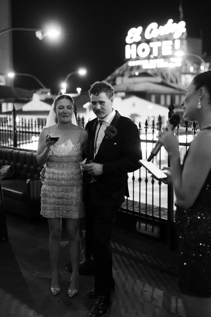 Bride and groom standing together as a guest gives a speech at their Commonwealth Las Vegas wedding reception.