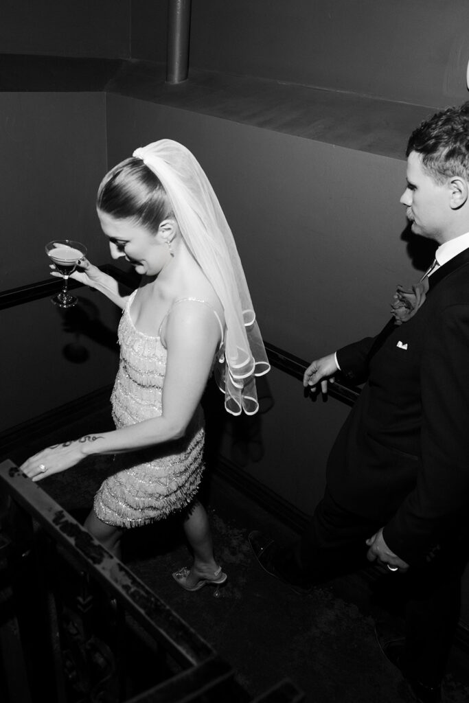Black and white photo of the bride walking down the stairs holding an espresso martini as the groom follows during the reception.