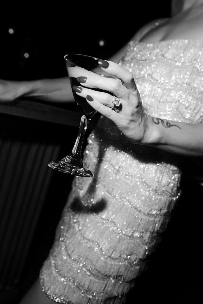 Close-up black and white image of the bride holding an espresso martini, highlighting her wedding ring and beaded dress.
