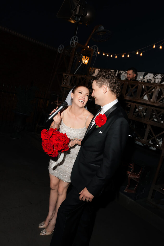 Bride laughing into a microphone while holding red roses beside her groom at their Commonwealth Las Vegas wedding reception.