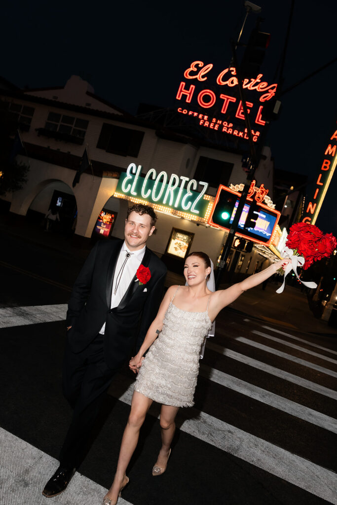 Newlyweds walking hand in hand through downtown Las Vegas at night with El Cortez neon signs in the background.