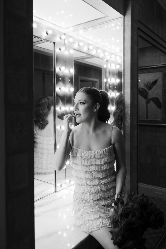 Black and white portrait of a bride applying lipstick in front of a lighted vanity mirror in a Las Vegas hotel suite.