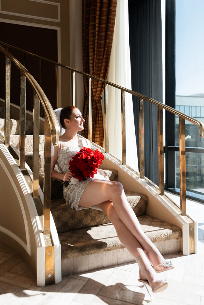 Bride sitting on the staircase at the Golden Nugget holding a red rose bouquet during getting ready photos.