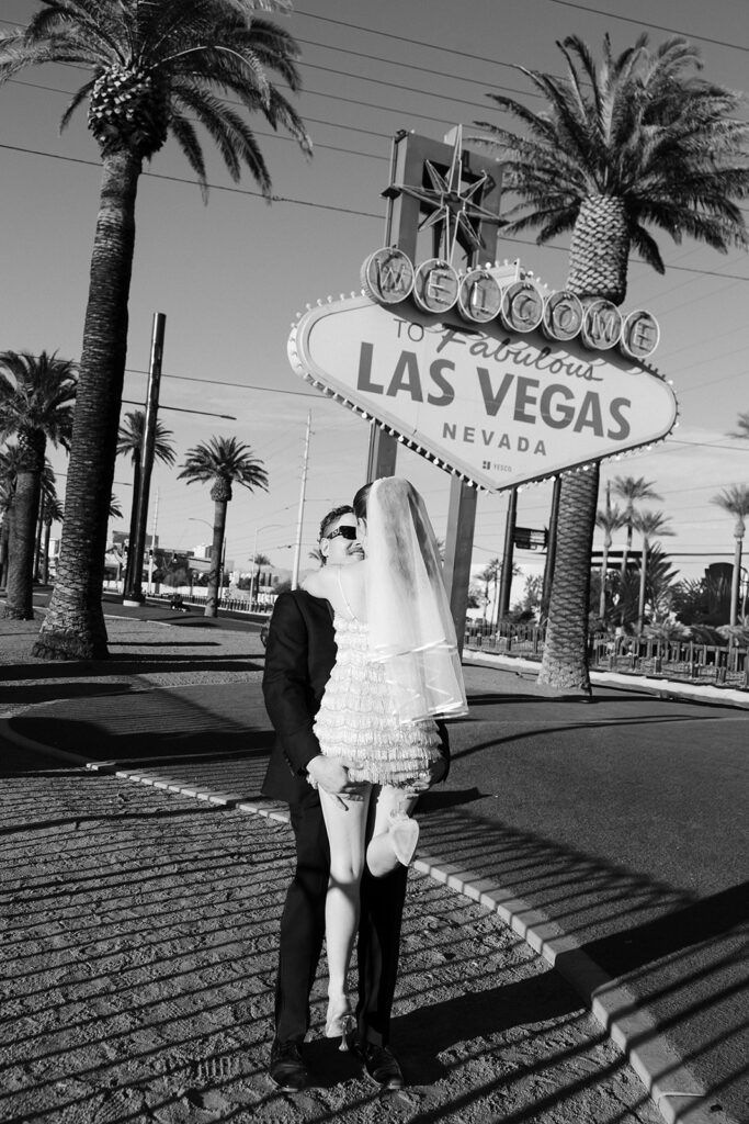 Bride and groom embracing beneath the Welcome to Fabulous Las Vegas sign in black and white.