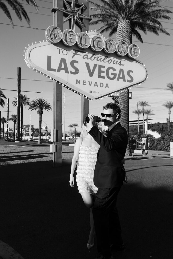 Black and white photo of a bride and groom drinking champagne in front of the Welcome to Fabulous Las Vegas sign.