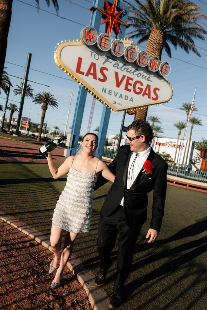 Bride and groom walking in front of the Welcome to Fabulous Las Vegas sign with a bottle of champagne.