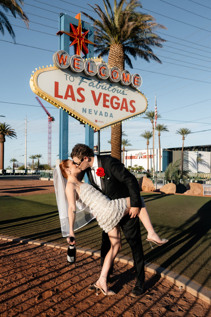 Bride and groom kissing in front of the Welcome to Fabulous Las Vegas sign with a bottle of champagne.