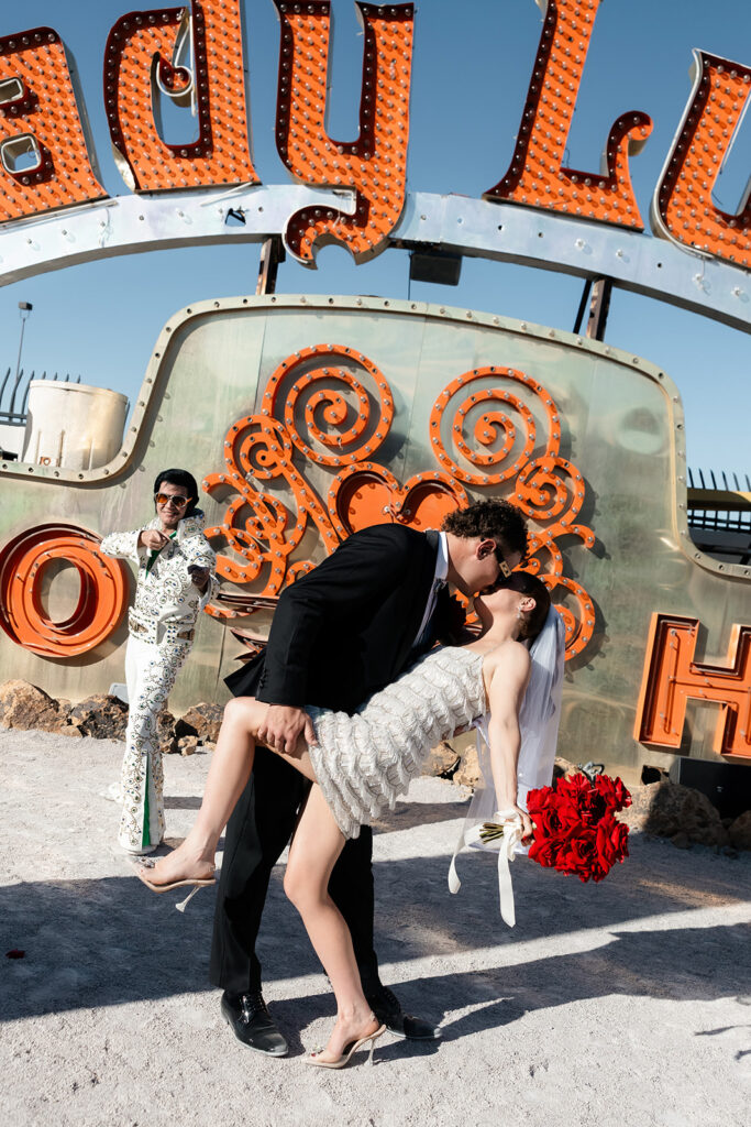 Bride and groom kissing with an Elvis officiant posing in the background during their Neon Museum wedding ceremony in Las Vegas.