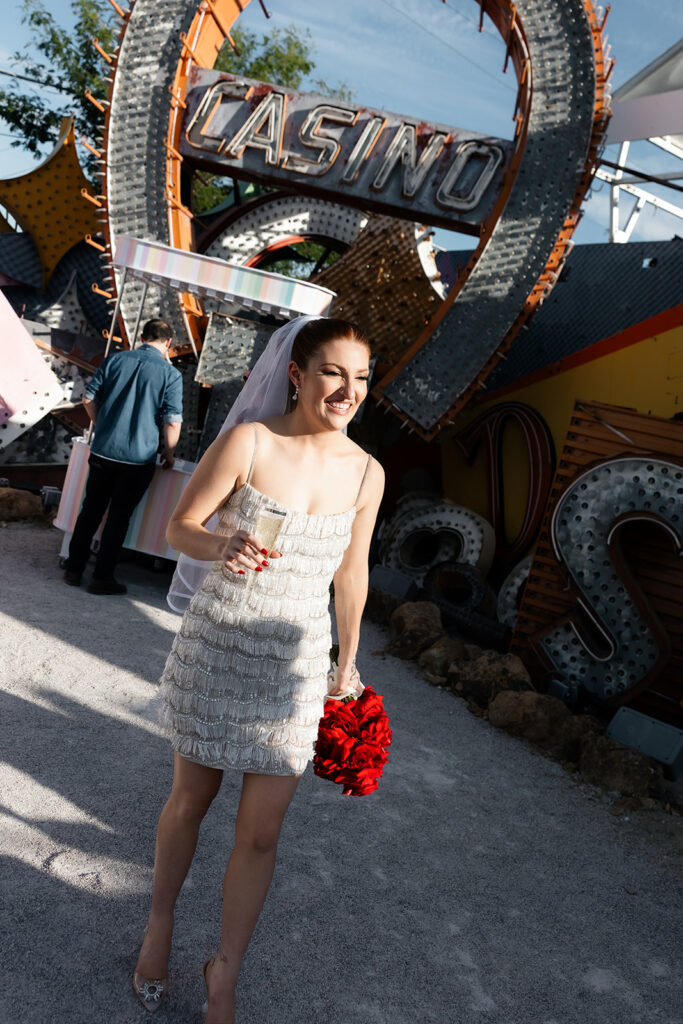 Bride smiling while holding a red rose bouquet beneath the Casino sign at the Neon Museum in Las Vegas.