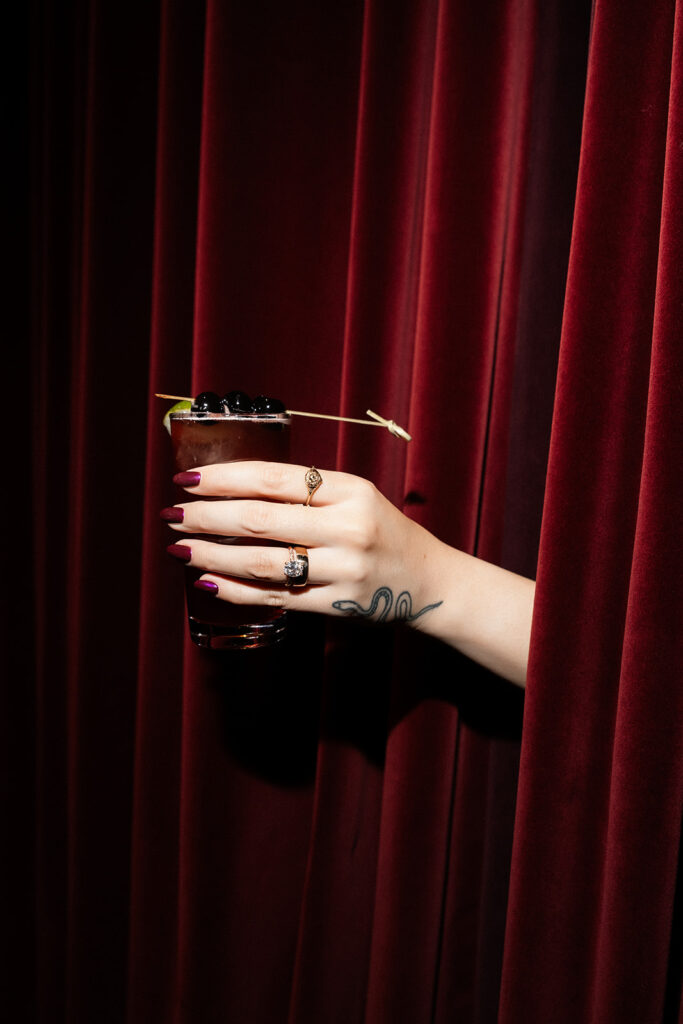 Bride holding a cocktail with olives in front of velvet curtains at the NoMad Library in Las Vegas.