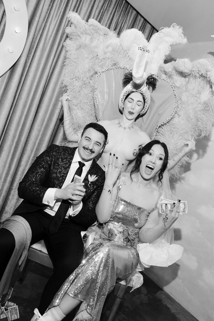 Black and white portrait of Surely the Showgirl standing behind a laughing couple at their Las Vegas micro wedding inside Sure Thing Chapel.