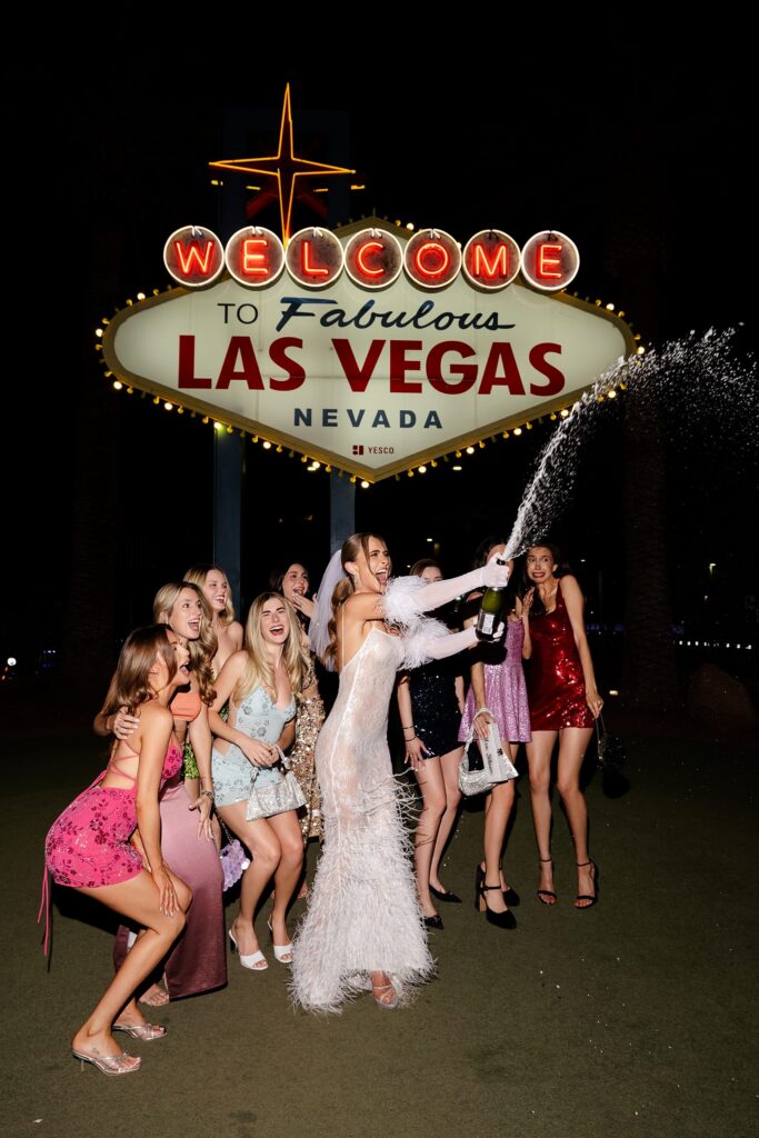 Bride and her friends popping champagne in front of the Welcoe4m to Fabulous Las Vegas sign at night. 