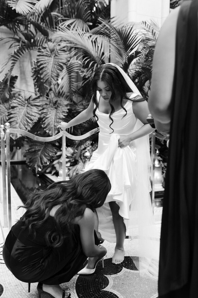 Candid black and white photo of a bride being helped into her heels.