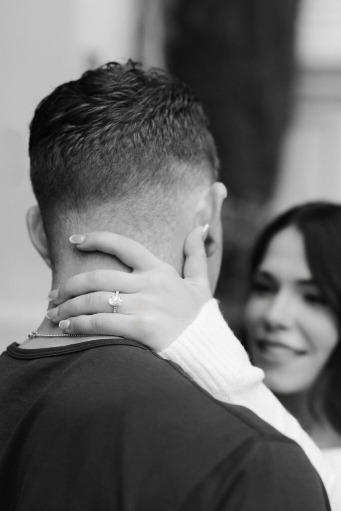 Black and white photo of a woman with her hands holding her fiancés neck and showing off her engagement ring.
