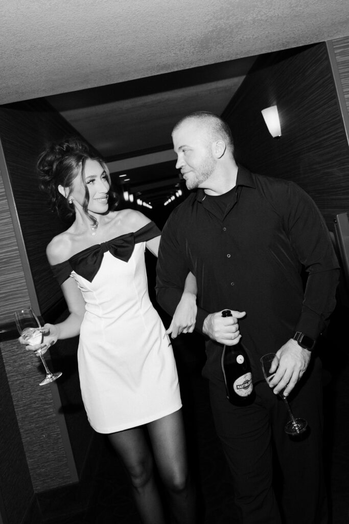 Couple holding champagne glasses and walking down a hotel hallway.