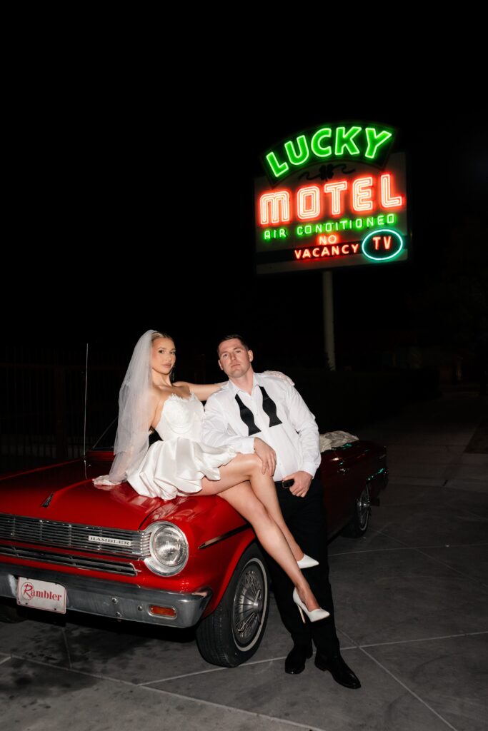 Couple posing with a red convertible in front of Lucky Motel outside of Sure Thing Chapel.