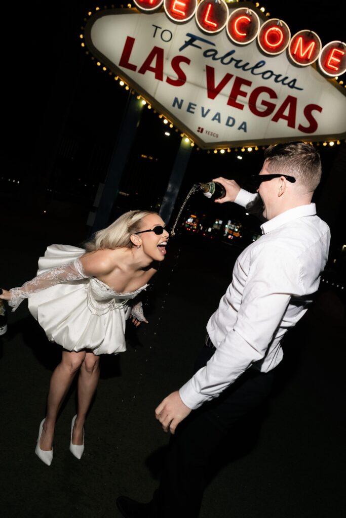 Man pouring champagne in his fiancés mouth in front of the Welcome to Fabulous Las Vegas sign. 