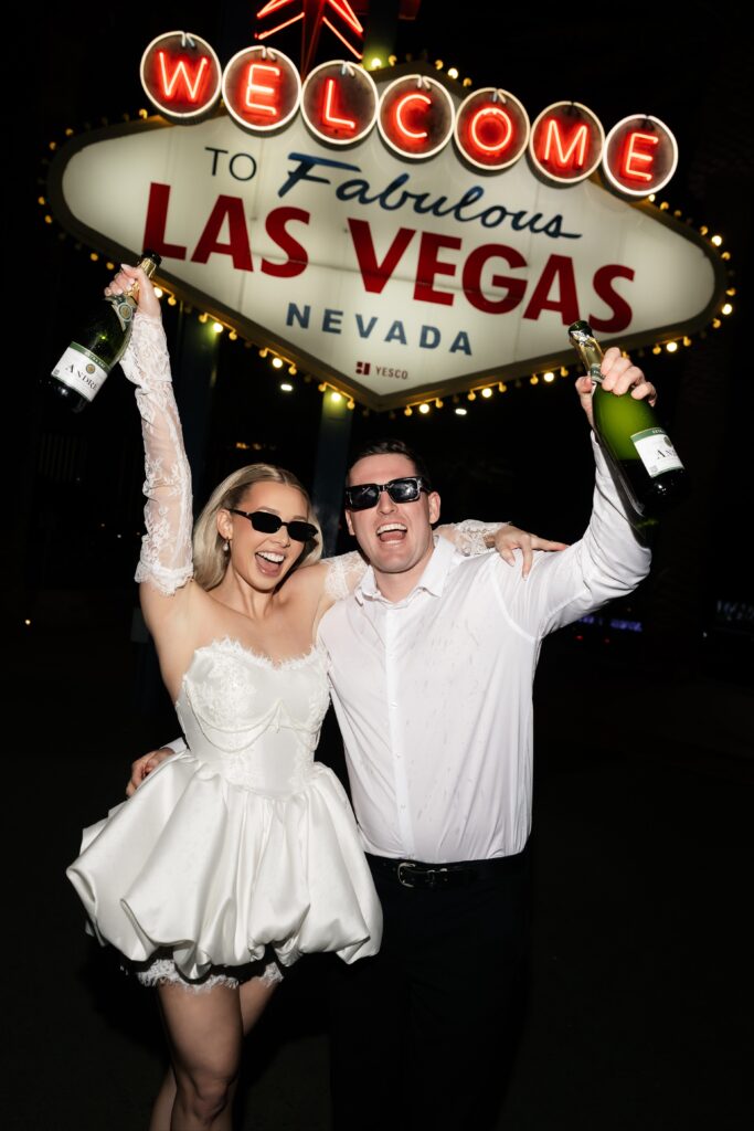 Couple holding up champagne bottles during their Las Vegas engagement photos in front of the Welcome to Fabulous Las Vegas sign. 