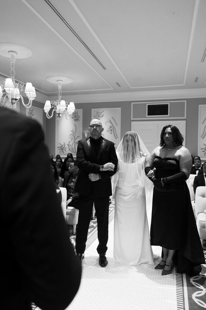 Black and white photo of the bride walking down the aisle with her mother and father for her Wynn Las Vegas Lavender Salon wedding ceremony.